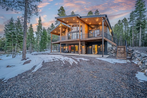Modern two-story cabin with wood and dark exterior, lit upper deck, surrounded by tall pine trees and snow at dusk.