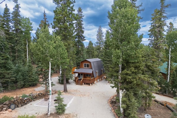 A-frame cabin with glowing windows and stone chimney surrounded by tall pine trees at dusk.