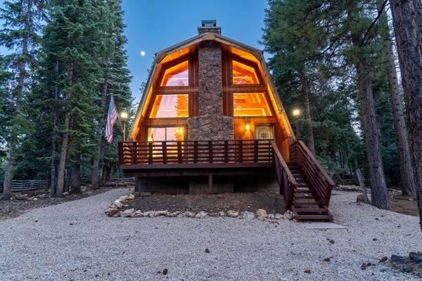A-frame cabin with glowing windows and stone chimney surrounded by tall pine trees at dusk.