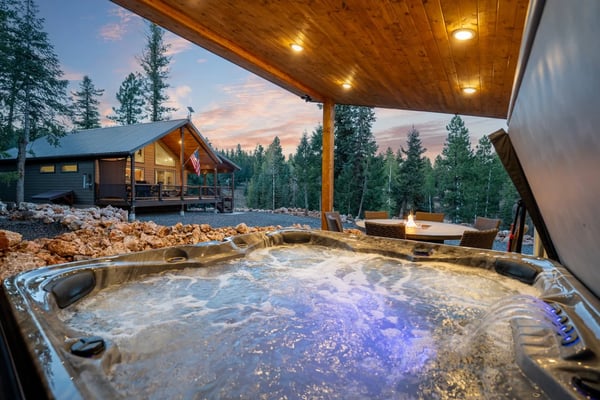 Hot tub under wooden pergola with forest cabin, fire pit, and pine trees at dusk