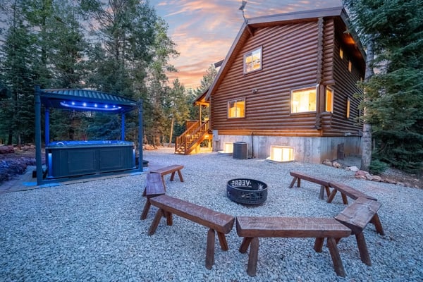 Log cabin at dusk with illuminated windows, fire pit, wooden benches, and blue-lit bar structure in forested setting.